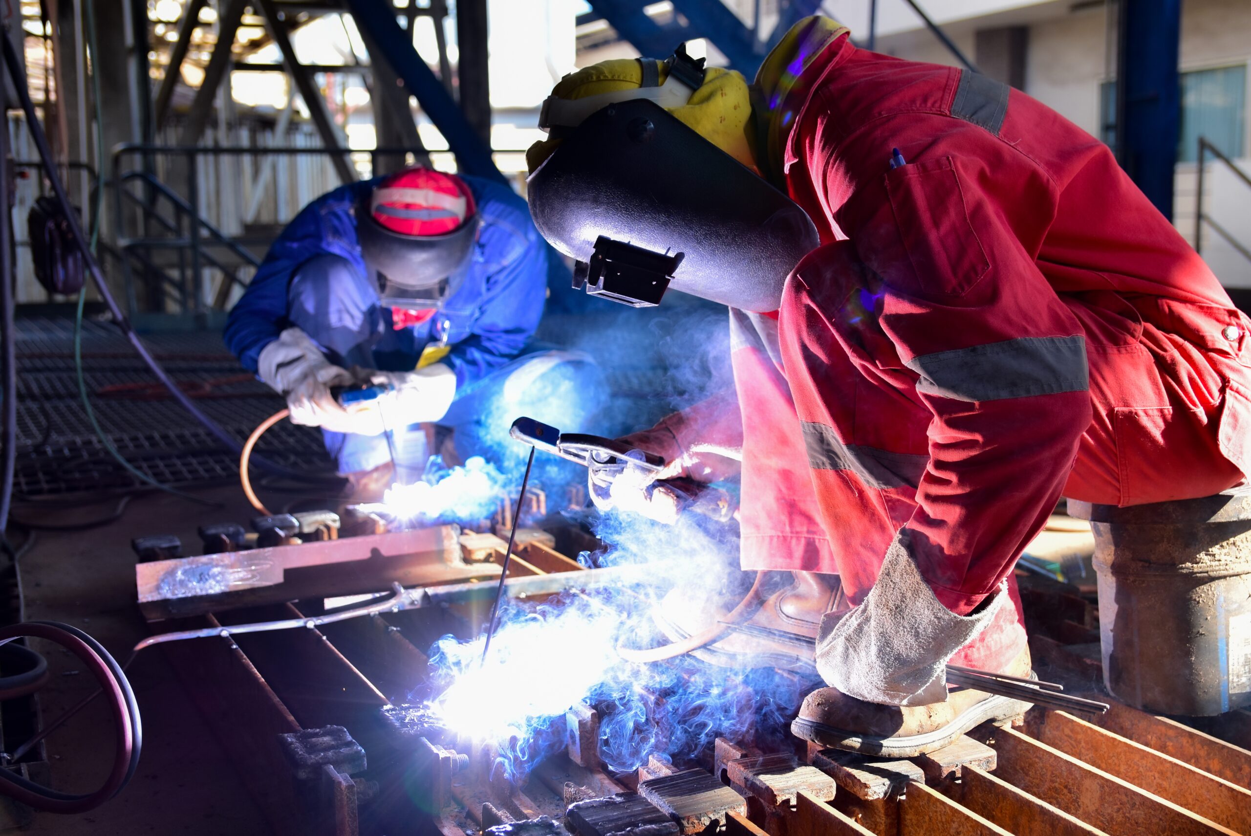 Two welder is assembling the workpiece by process shielded metal arc welding (SMAW). Welder in red uniform, safety shoes, leather gloves, welding mask. He is sitting and welding.